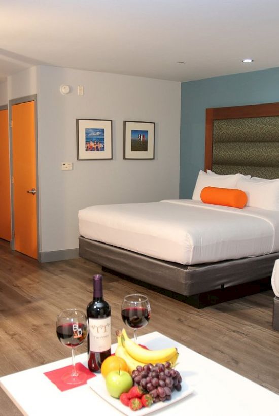A modern hotel room with two beds, a desk, chairs, wine, and a plate of fruits on a table. Bright and clean with contemporary decor.